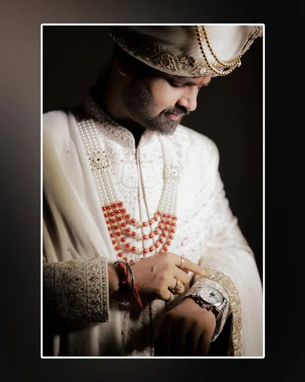 A close-up, sophisticated portrait of the groom checking his watch. The dramatic lighting highlights the rich details of his white sherwani and the intensity of the moment.