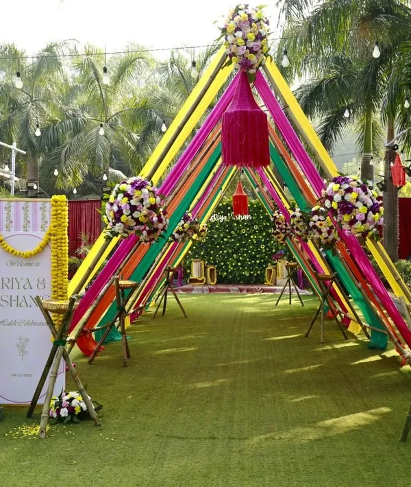 A perfect entrance for a vibrant Haldi ceremony. The welcome sign for Riya and Ishaan stands amidst a tunnel of colorful ribbons and marigolds, setting a festive tone right from the start.