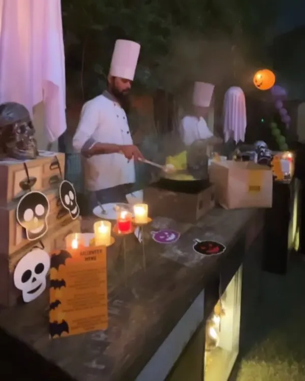 A chef at a live cooking station during a Halloween party. The station is decorated with spooky skulls, skeletons, and candles, fully immersing the cooking experience into the theme.