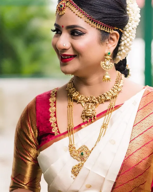 A classic South Indian bridal portrait. This bride is wearing a traditional off-white and red silk saree, complemented by antique gold temple jewellery. Her makeup features a sharp winged liner, a red bindi, and a bold red lip.