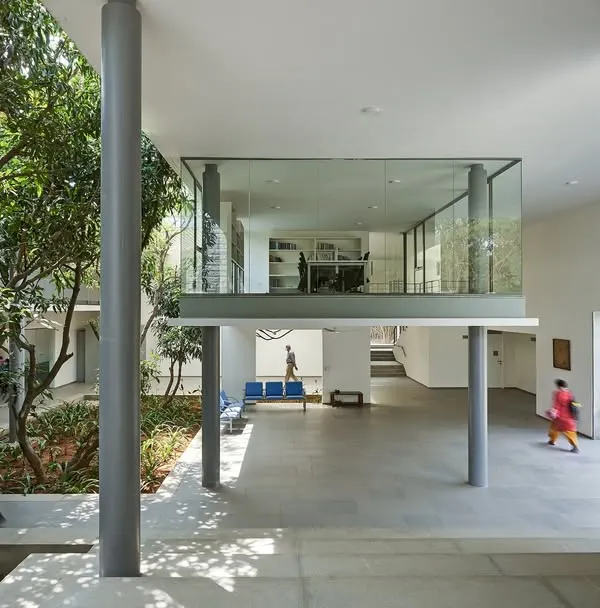 A floating glass box containing a discussion room overlooks a landscaped court at the Karunashraya Institute for Palliative Care (KIPCER). The design creates transparent, light-filled spaces for learning and collaboration.