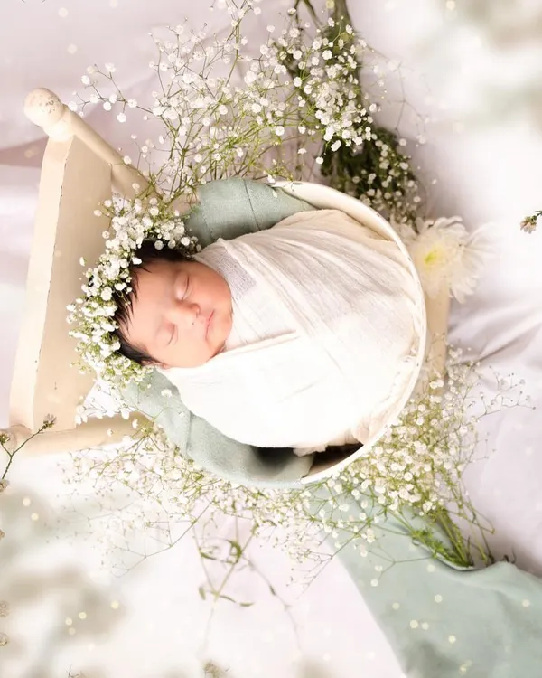 Wrapped snugly in white fabric and adorned with a floral crown of baby's breath, a newborn sleeps peacefully. This image highlights the delicate details and angelic calm of a professionally styled newborn session.