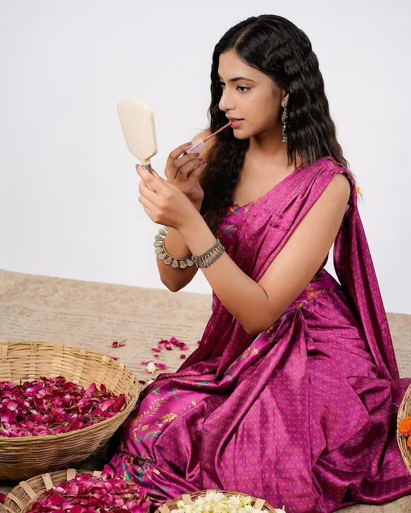 A beauty shoot for a makeup brand, showing a model applying lip gloss while looking in a handheld mirror. The setting with rose petals adds a touch of natural elegance to the product shot.