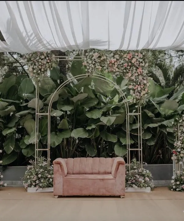 This minimalist and modern wedding stage is perfect for a chic garden ceremony. A simple gold geometric arch is adorned with delicate pink and white florals, set against a backdrop of lush natural greenery, with a blush pink sofa for the couple.