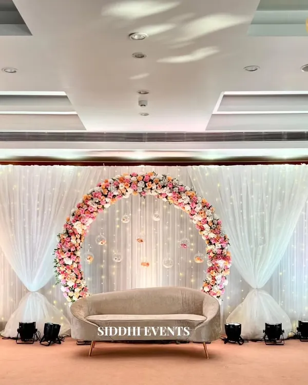 A perfect circle of pastel roses and hydrangeas frames a chic beige sofa, creating a stunningly romantic and elegant stage for a ring ceremony. The fairy light curtain adds a touch of sparkle to the backdrop.
