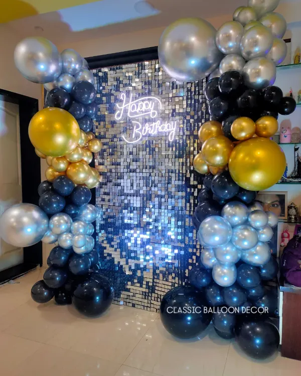 A stunning silver shimmer wall framed by black, gold, and silver balloon pillars. The 'Happy Birthday' neon sign adds a modern, celebratory glow.