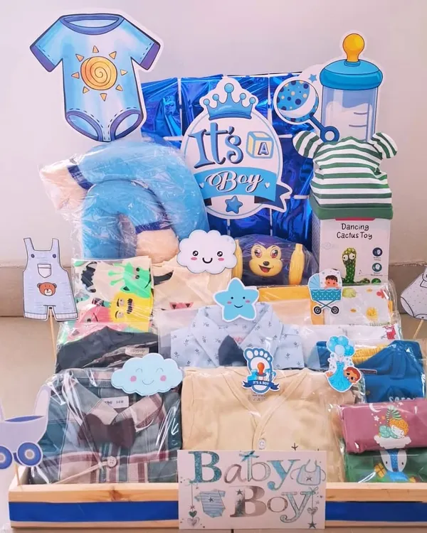 An 'It's a Boy' hamper packed with a variety of clothes, a dancing cactus toy, and other essentials. The blue theme is carried throughout, from the decorations to the baby items.
