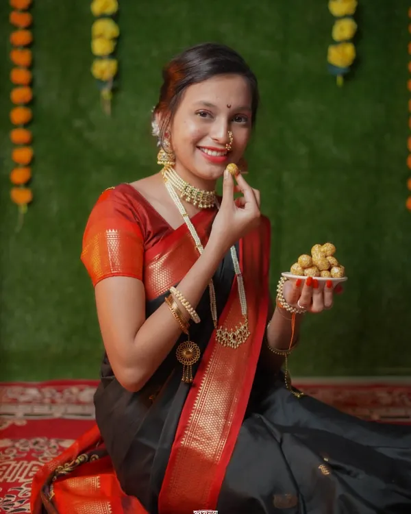 A classic Makar Sankrant portrait. The model is dressed in a traditional black and red saree, holding a plate of 'tilgul ladoo' against a festive backdrop of marigold flowers.