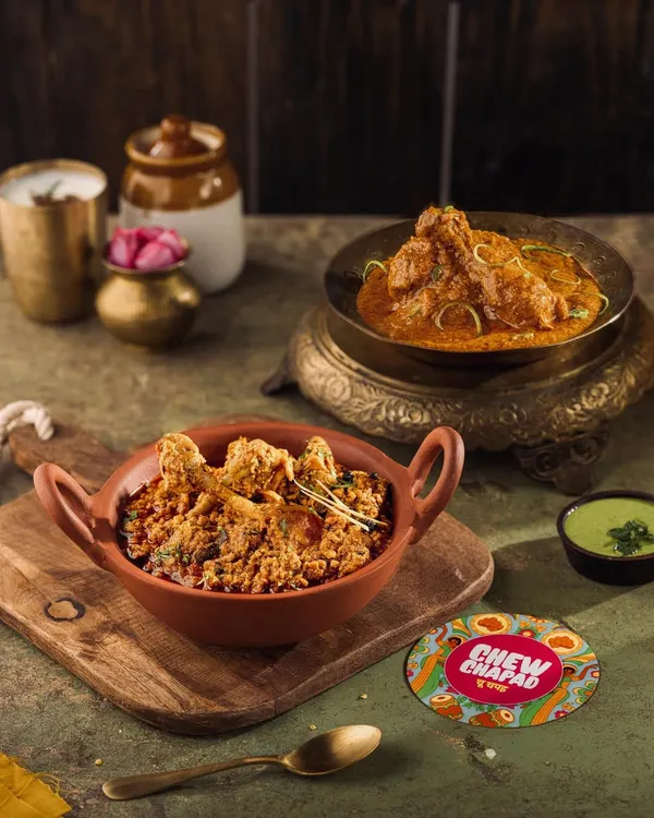A rustic composition featuring Chicken Curry and Mutton Kheema for Chew Chapad. The use of traditional earthenware and brass bowls against a dark background enhances the rich, home-cooked feel of the dishes.