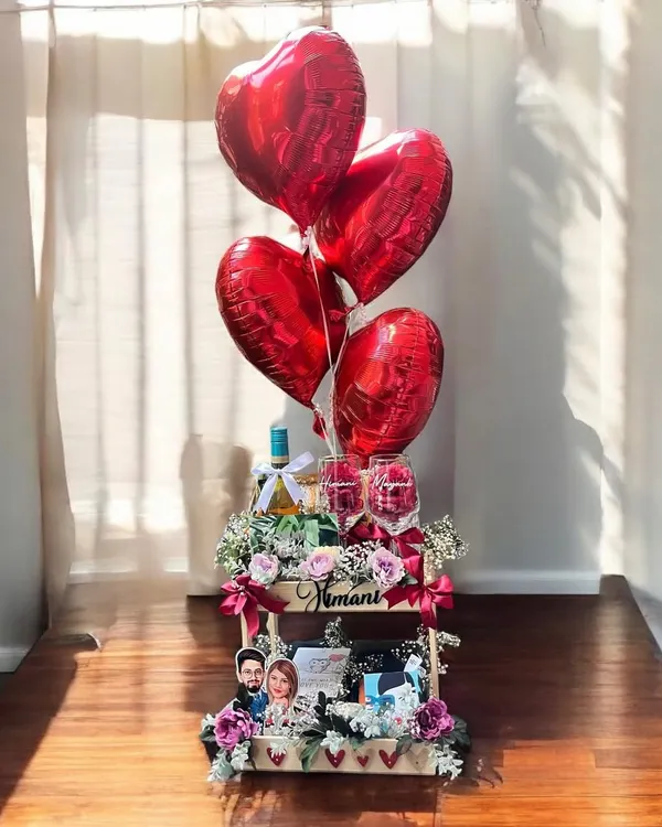 I designed this romantic anniversary hamper to be a showstopper, featuring a large wooden crate filled with flowers, personalized photo frames, wine, and elegant glasses, all topped with heart-shaped red foil balloons.