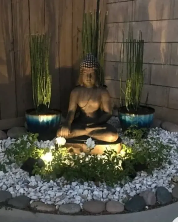 A serene Zen corner designed for evening relaxation. This setup features a meditating Buddha statue as the focal point, surrounded by white pebbles, uplighting, and minimalist plants for a tranquil ambiance.