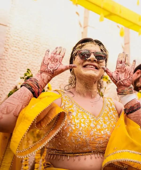 Nothing beats the pure, unscripted fun of a Haldi ceremony. This bride, covered in turmeric paste and sporting sunglasses, shows off her mehndi with a huge smile. We capture these happy, candid moments.