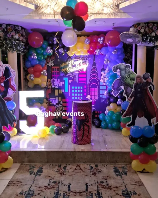 A full view of a superhero themed birthday party setup. This decoration features character cutouts of Thor, Batman, and Spiderman, a large marquee number '5', and a custom city backdrop with a neon 'Happy Birthday' sign.