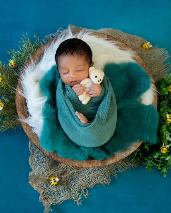 A top-down view of a baby boy swaddled in a teal wrap, holding a tiny bear. The setup includes greenery and small bee props against a rich blue backdrop, creating a nature-themed scene.