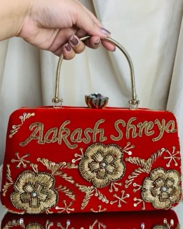 A vibrant red velvet clutch, personalized with the names 'Aakash' and 'Shreya' in beautiful golden script. The floral embroidery adds a touch of elegance to this custom bridal accessory.