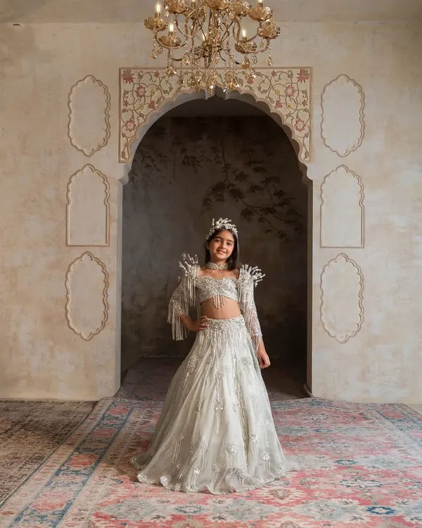 We design outfits that make your little girl feel like the princess she is. This breathtaking antique green lehenga, with its intricate pearl and tassel work on the sleeves and choker, is a perfect example of our handcrafted luxury for those truly special occasions.