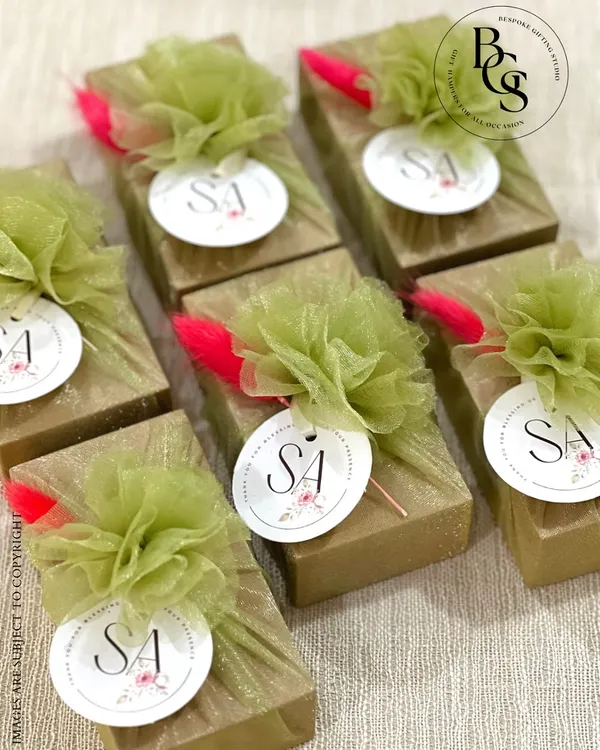 Charming return favors for a special event. These small gifts are wrapped in gold glitter paper and adorned with a fluffy green tulle pom-pom, a pop of pink, and a custom monogrammed tag.