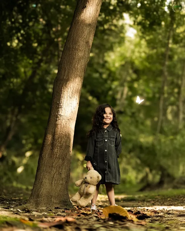 A magical moment of childhood innocence in the woods. This little girl's bright smile, her teddy bear companion, and a fluttering butterfly create a truly enchanting outdoor portrait.