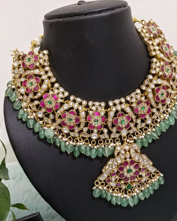 A breathtakingly beautiful Jadau necklace set with intricate floral motifs in pink and green, accented with uncut polki stones and natural strawberry beads. This is paired with matching chaandbaali earrings for a complete bridal look.