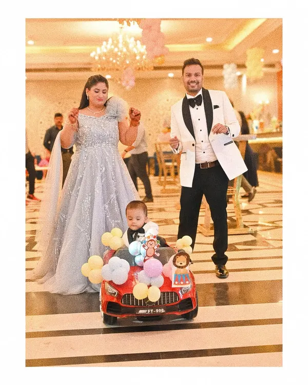 The birthday boy makes a memorable entrance in a toy car decorated with balloons, as his elegantly dressed parents dance alongside him. This shot captures the fun and festive atmosphere of the celebration.