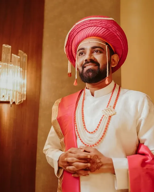 A classic portrait of a Marathi groom, looking dapper in his traditional wedding attire. The pink turban (pheta) and beaded jewelry add a touch of regal elegance.