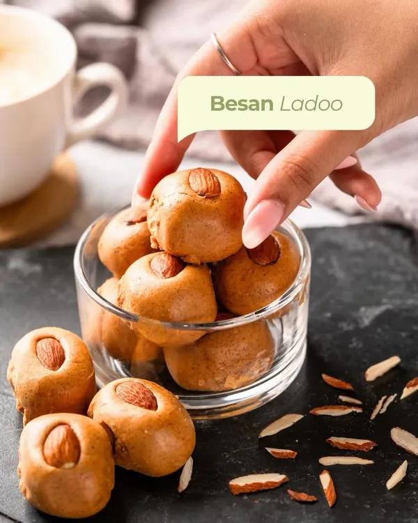A hand reaches for a classic Besan Laddoo from a glass bowl. Made with slow-roasted besan and topped with an almond, it's the perfect comforting sweet to have with your evening chai.