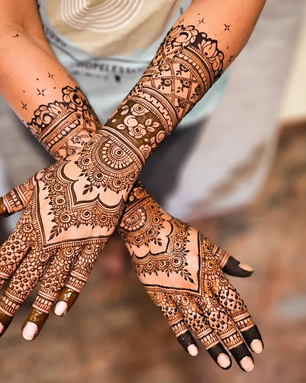 A classic and dense bridal mehendi design. The palms are covered in a symmetrical pattern featuring a central heart-shaped motif, surrounded by detailed floral and geometric work that extends up the forearm.