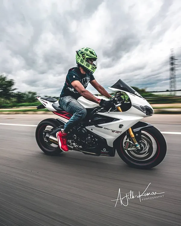 A full view of a rider on a white Triumph Daytona sports bike, captured with a panning motion blur technique. The sharp subject against the streaked background perfectly conveys the speed of the ride.