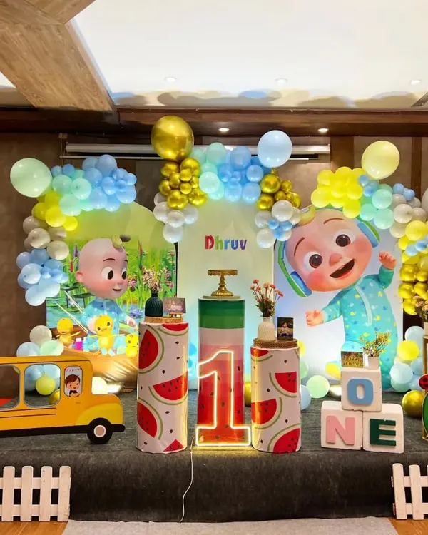 A vibrant Cocomelon themed first birthday party for Dhruv. This large stage setup includes multiple character cutouts, a watermelon-themed cake stand, a light-up number "1," and a yellow school bus prop.