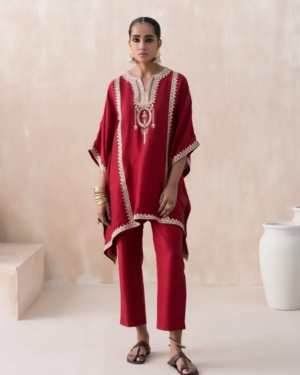 A clean, full-length lookbook image styled for Saisha's 'Firdaus' collection. The focus is on the sharp silhouette of this red kaftan set, with minimal props and a neutral background to make the intricate embroidery and vibrant color stand out.
