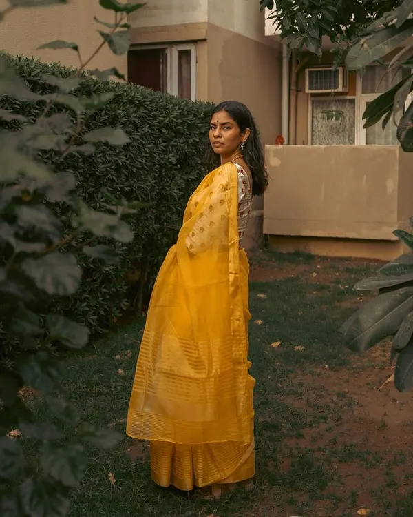 A full-length portrait of a woman in a yellow saree, framed by lush garden foliage. This composition highlights the elegance of the drape in a natural, serene environment.