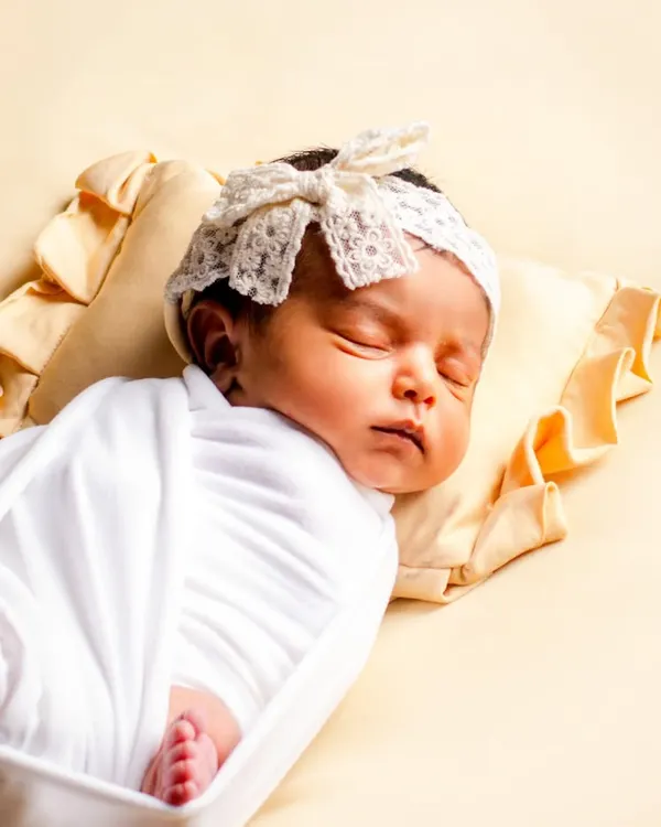 This is a classic newborn portrait focusing on a safely swaddled baby. The simple white wrap and lace headband highlight the baby's peaceful expression and delicate features against a soft yellow backdrop.