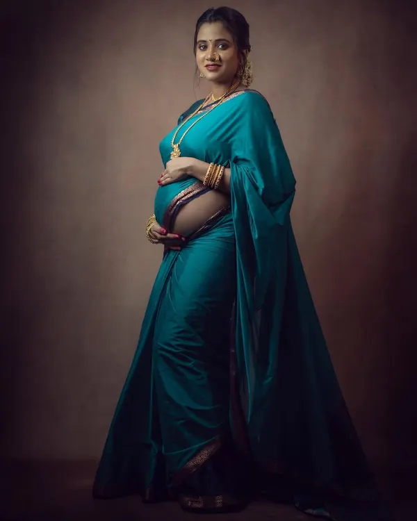 A full-length portrait showcasing the grace of a mother-to-be in a beautiful teal saree. The studio lighting is carefully set to highlight the texture of the fabric and the gentle curve of the baby bump.