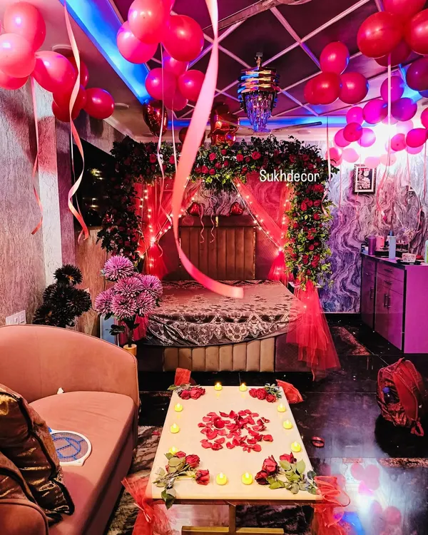 This is a wide view of a complete romantic room decoration. The setup includes a bed canopy of red roses, fairy lights, red balloons on the ceiling, and a table set with candles and rose petals for a memorable surprise.