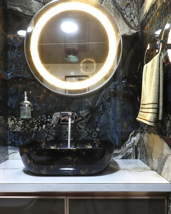 A luxurious powder room featuring a statement black and gold vessel sink. The dramatic black marble wall and backlit circular mirror create a bold and sophisticated atmosphere.