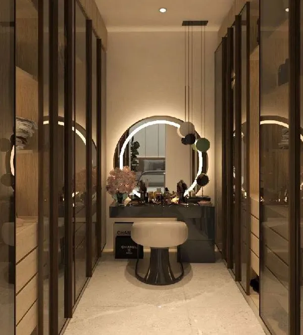A luxurious walk-in closet featuring floor-to-ceiling glass-door wardrobes and a central vanity. The circular backlit mirror and elegant stool create a glamorous, boutique-like dressing experience.