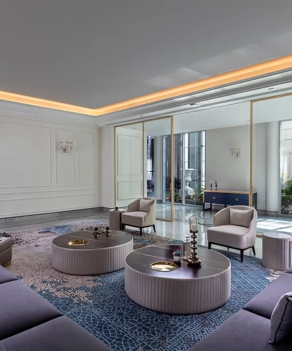 In The Grand Alabaster, soft light from the courtyard fills this formal living room, creating a warm and still atmosphere. The combination of a plush sectional sofa, a patterned rug, and classic wall paneling gives the space a sense of calm, comfort, and quiet luxury.