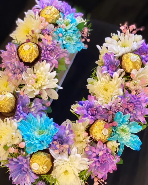 A vibrant and colorful monogram arrangement. This letter 'U' is crafted with a mix of purple, blue, and white chrysanthemums and dotted with Ferrero Rocher chocolates for an extra touch of luxury.