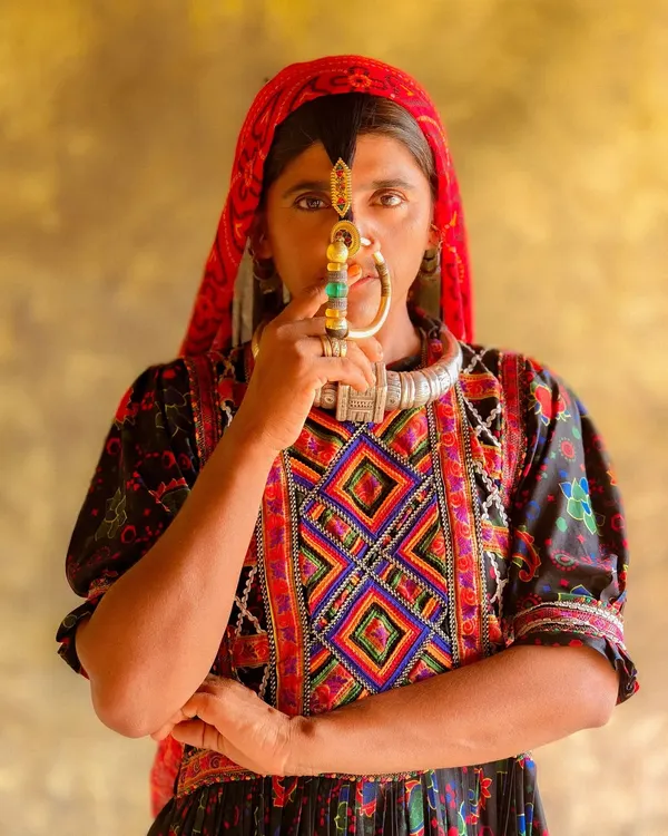 A powerful portrait of a Dhaneta Jat woman from Kutch, her face partially obscured by the large, traditional nose ring she holds. Her direct gaze is captivating and full of strength.