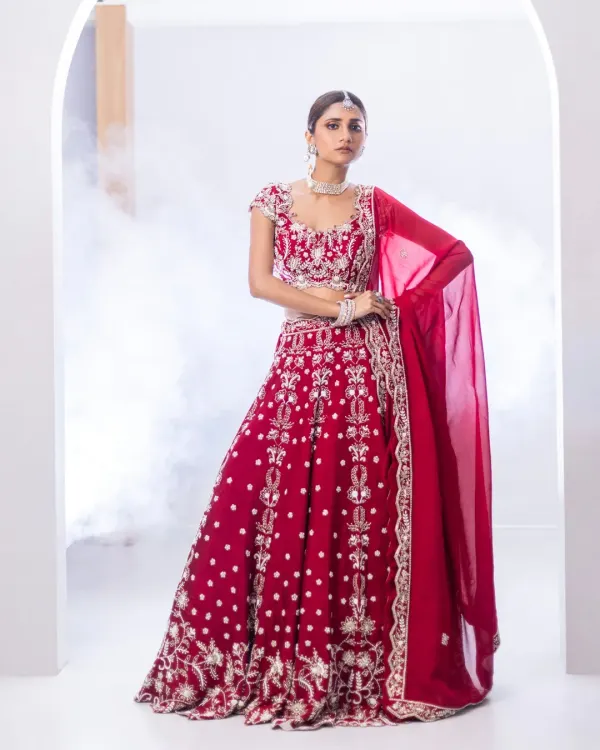 A traditional red bridal lehenga, reimagined for the modern bride. The full-volume skirt and intricately embroidered blouse, featuring sequins and metallic threadwork, create a look that is both classic and breathtaking.