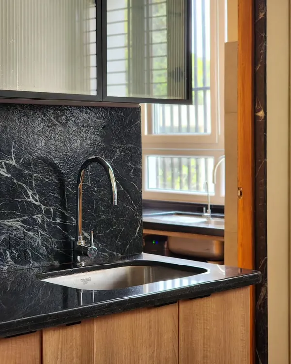 A close-up of a kitchen sink area, showcasing the rich texture of the black marble backsplash against the warm wooden cabinetry. The fluted glass of the overhead cabinet adds a layer of translucency and refinement.