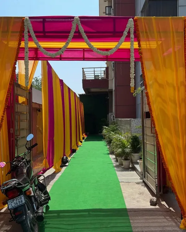 A vibrant entrance for a daytime event, created with yellow and pink drapes and marigold garlands over a green carpet walkway.