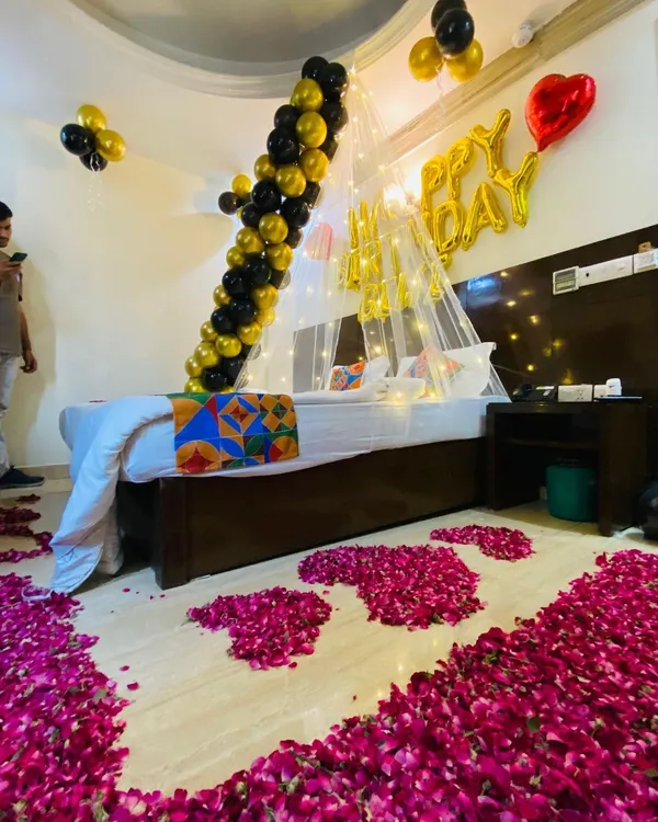 A luxurious birthday room decoration featuring a path of rose petals leading to the bed. The decor includes a canopy with fairy lights, a black and gold balloon garland, and "Happy Birthday" foil balloons.