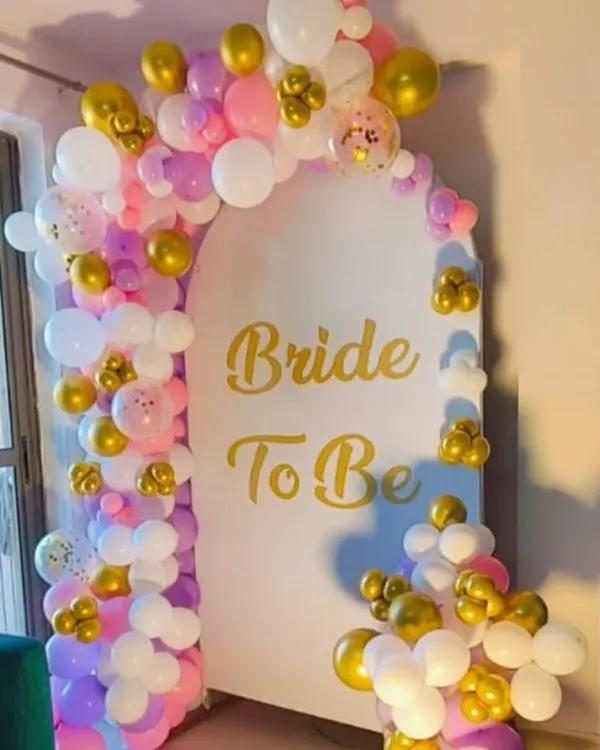 A lovely "Bride to Be" backdrop for a home party. This setup features a white arched panel with gold lettering, framed by a colorful balloon garland in shades of pink, purple, white, and gold.
