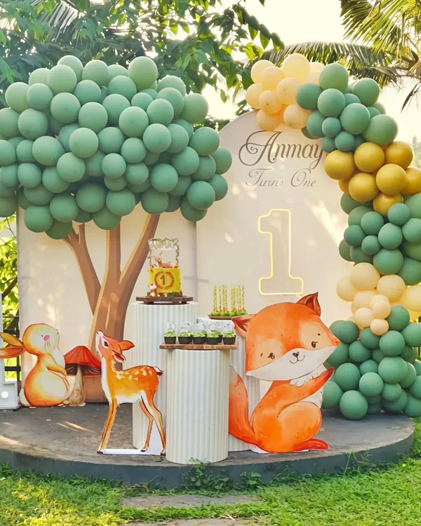 An enchanting outdoor woodland animal theme for a first birthday. This setup features a custom balloon tree, large character cutouts of a fox and deer, and multiple pedestals for the cake and treats, all set on a circular stage.