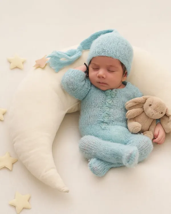A baby in a cozy blue sleeper and hat cuddles a bunny while sleeping on a soft crescent moon pillow, surrounded by stars.