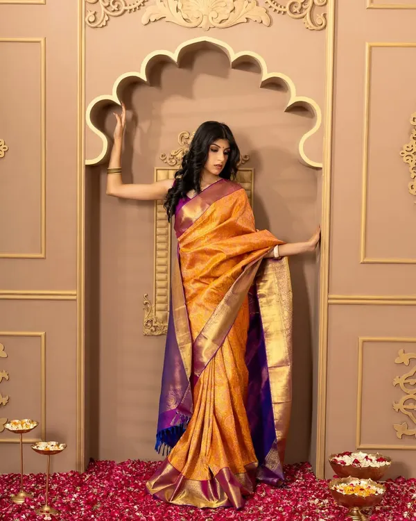A full-length shot of a model in a vibrant orange and purple silk saree from Kumaran Silks. The grand, traditional backdrop complements the festive and formal nature of the attire.