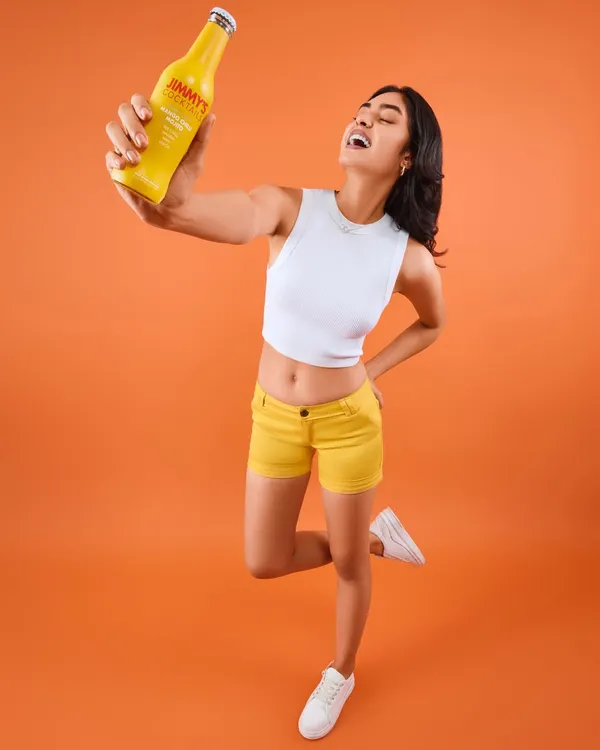 A full-length shot from the Jimmy's Cocktails campaign, capturing a moment of pure joy. The styling is bright and energetic, with the model in yellow shorts and a white crop top that perfectly matches the brand's vibrant and fun-loving identity.