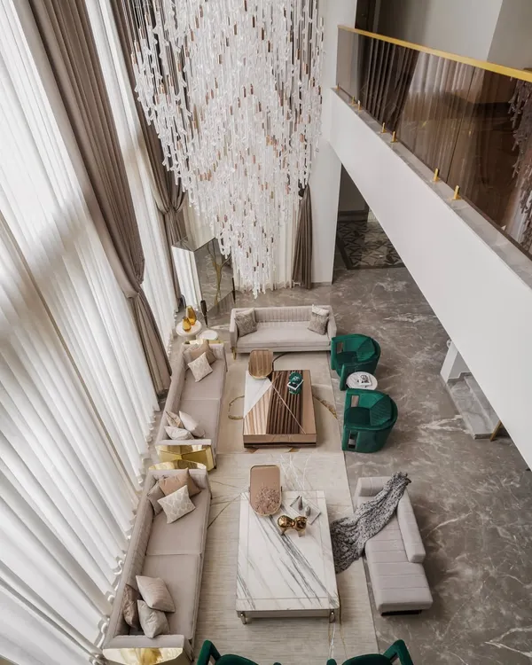 An aerial view of a double-height living room, showcasing the grandeur and openness we designed into the space. The layout is planned for easy conversation, with distinct seating zones defined by emerald green chairs and neutral sofas, all anchored by a magnificent crystal chandelier.