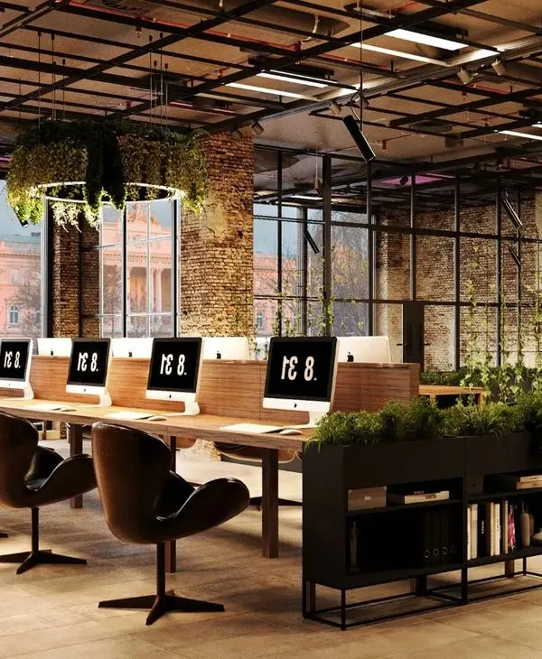 This industrial-style office design uses exposed brick, high ceilings, and abundant natural light to create an inspiring, collaborative workspace. The inclusion of extensive greenery and a central hanging plant feature brings a biophilic element that boosts well-being and creativity.